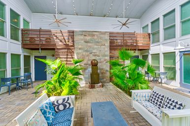 Sunlit covered courtyard patio with lush potted palms, a central stone fountain, wooden slatted balcony, hanging ceiling fans and blue lounge seating