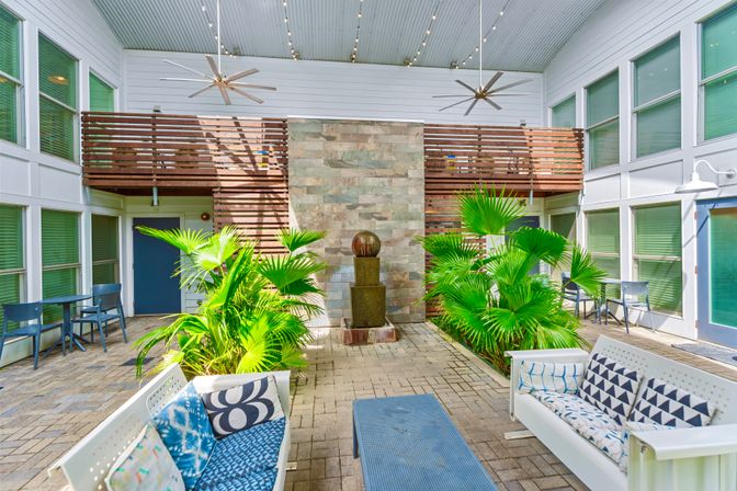 Sunlit covered courtyard patio with lush potted palms, a central stone fountain, wooden slatted balcony, hanging ceiling fans and blue lounge seating