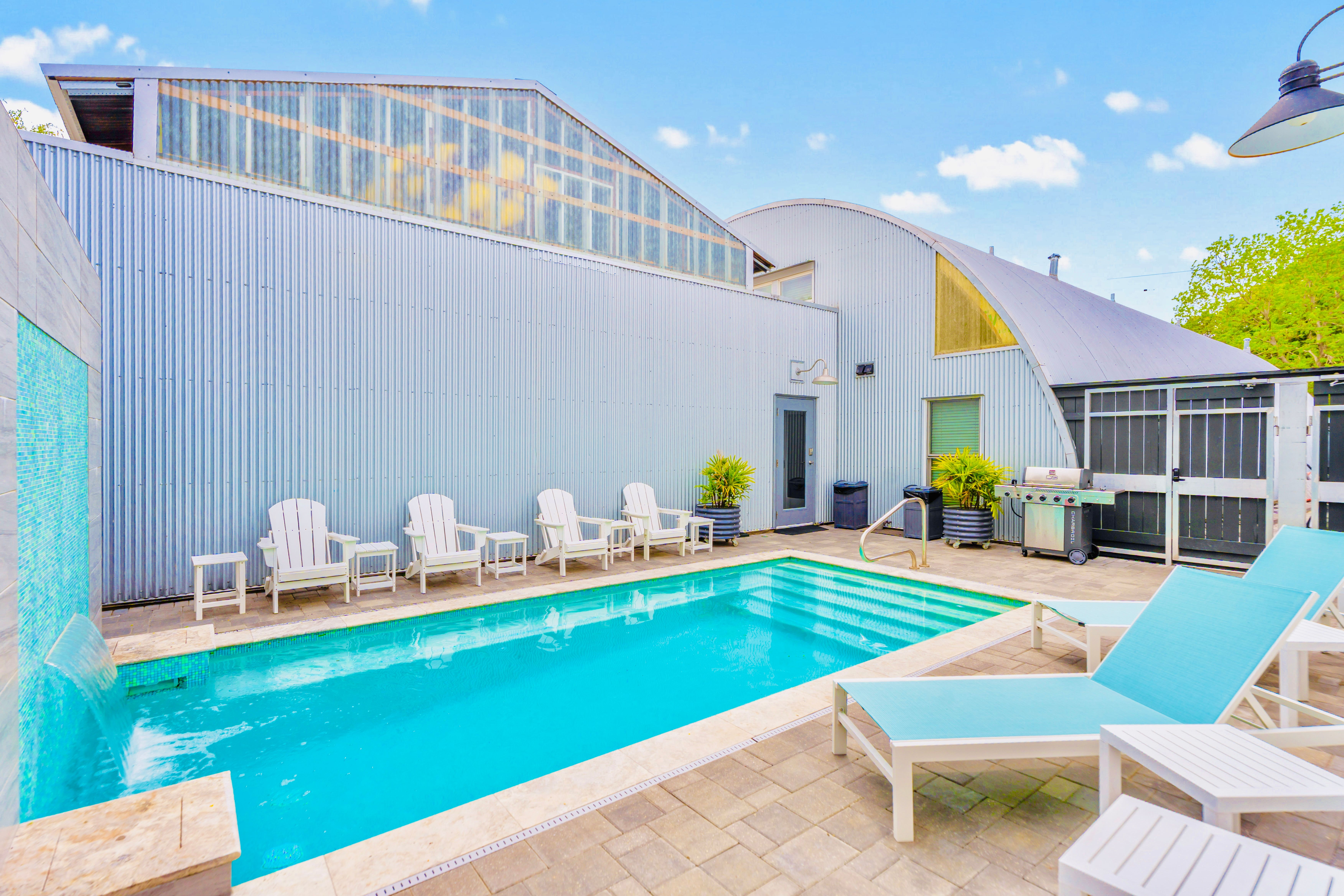 Bright outdoor pool in an industrial‑chic courtyard with corrugated metal buildings, turquoise lounge chairs, white Adirondack seats, potted plants and a stainless grill under a clear blue sky