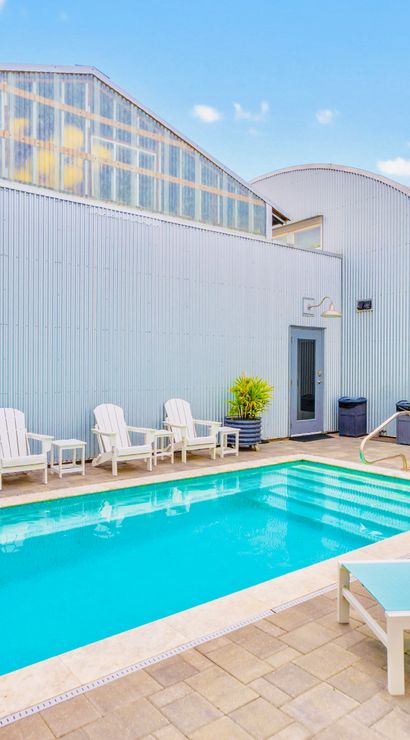 Bright outdoor pool in an industrial‑chic courtyard with corrugated metal buildings, turquoise lounge chairs, white Adirondack seats, potted plants and a stainless grill under a clear blue sky