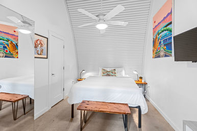 Bright modern minimalist bedroom with white bedding beneath an arched corrugated metal ceiling, white ceiling fan, wooden bedside tables with lamps, bench at foot of bed, colorful wall art and wall-mounted TV.