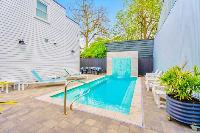 Sleek backyard lap pool with turquoise water and mosaic waterfall feature, flanked by white siding house, blue lounge chairs, outdoor dining table, potted plants and tall trees