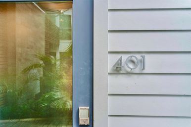 Sleek blue-framed glass front door with keypad lock, green palm reflections in the glass, and silver house numbers "401" mounted on white horizontal siding — modern home entrance.