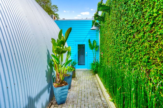 Narrow paved courtyard leading to a bright turquoise door, flanked by a corrugated metal wall on one side, a dense ivy-covered wall with an outdoor wall lamp on the other, and tall potted tropical plants along the walkway.