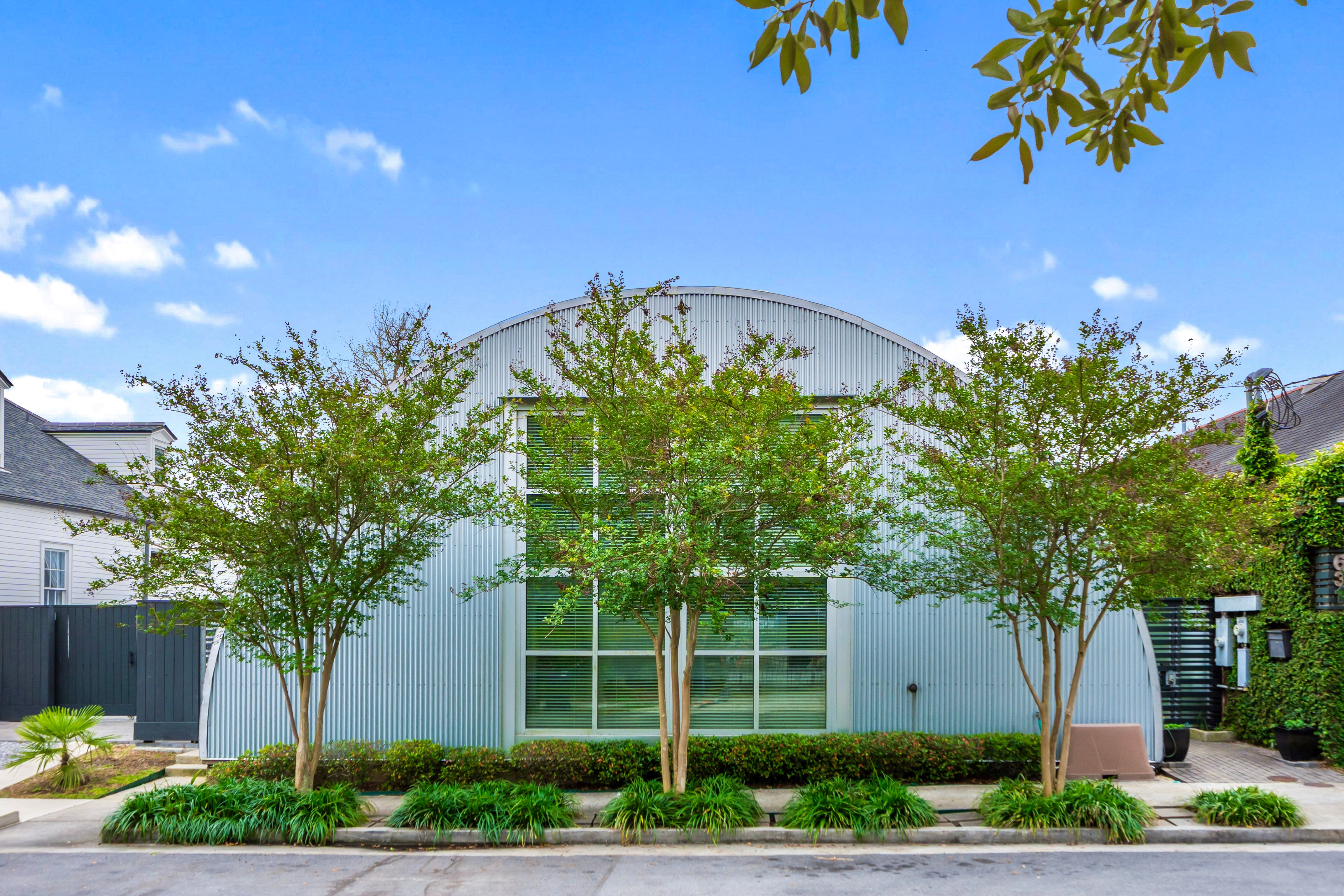 Sleek arched corrugated-metal building facade with large street-level windows, young trees and manicured shrubs lining the sidewalk under a bright blue sky