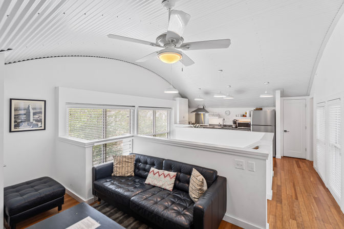 Bright modern loft apartment interior with white corrugated vaulted ceiling and ceiling fan, black tufted leather sofa with patterned pillows, open kitchen with pendant lights, stainless fridge and hardwood floors.