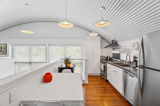 Bright modern loft kitchen with arched corrugated ceiling, three hanging pendant lights, white cabinets, stainless-steel appliances, wood floors and a table with a patterned cloth and red decorative vase.