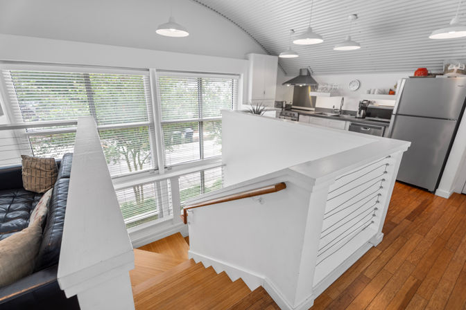 Bright modern loft with white staircase and wood floors overlooking an open-plan kitchen with stainless steel appliances, pendant lights, and large windows with horizontal blinds.