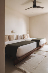 Minimalist modern twin bedroom with low wooden platform beds, white linens, olive-green throws, textured beige pillows, cream walls and a black ceiling fan.