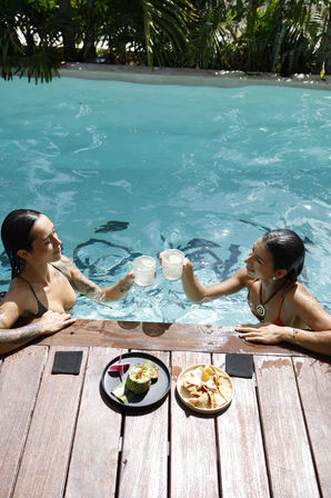 Two people clinking drinks in a turquoise tropical pool beside a wooden deck with guacamole and tortilla chips.