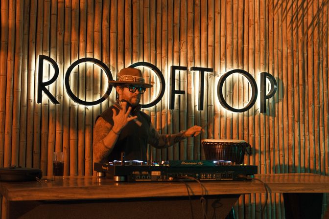 DJ spinning at a rooftop bar at night in front of a backlit sign on a bamboo wall, wearing a hat and glasses while mixing on DJ equipment under warm orange lighting.