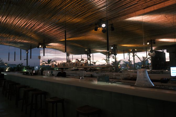 Rooftop bar at sunset with a long counter, warm wooden slatted ceiling and hanging pendant lights, rows of bottles and tropical plants silhouetted against a dusky skyline.