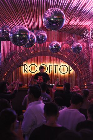 Rooftop bar scene with mirrored disco balls, DJ behind a bamboo arch, purple-pink neon lighting, tropical plants and a dancing crowd