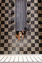Overhead view of an indoor black-and-white checkerboard tile floor with a long dark wooden communal table, round stools lining both sides, and two people seated at one end.
