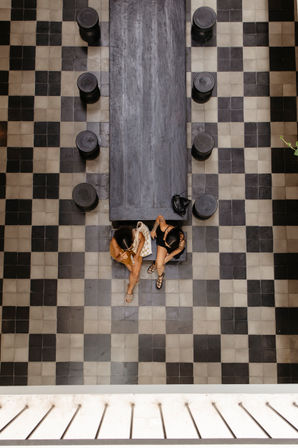 Overhead view of an indoor black-and-white checkerboard tile floor with a long dark wooden communal table, round stools lining both sides, and two people seated at one end.