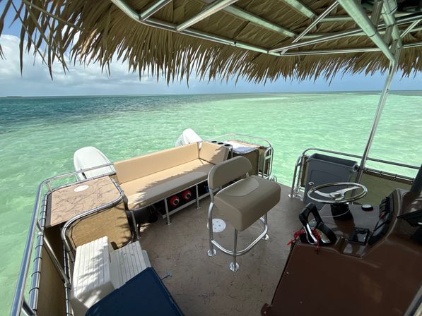 Pontoon boat with tan bench seating, captain’s chair and helm under a thatched palapa, floating over clear turquoise tropical lagoon water on a sunny day.
