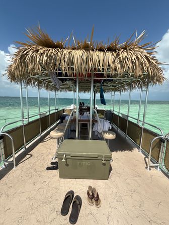 Pontoon boat with a palm-thatched tiki roof floating over clear turquoise tropical water, beige deck with a large cooler and two pairs of flip-flops, metal railings and sunny blue sky.