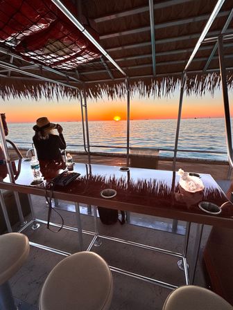 Hat-wearing person photographing a vivid orange sunset over calm coastal ocean from a thatched-roof pontoon boat, with a glossy bar table and stools reflecting the sky