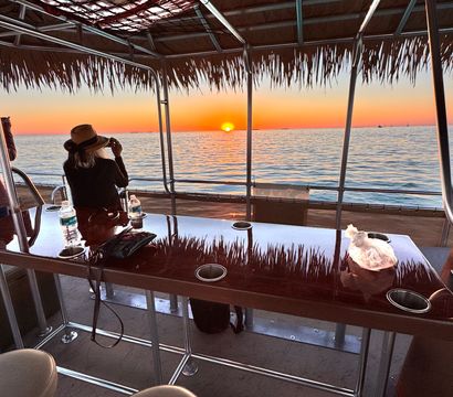 Hat-wearing person photographing a vivid orange sunset over calm coastal ocean from a thatched-roof pontoon boat, with a glossy bar table and stools reflecting the sky