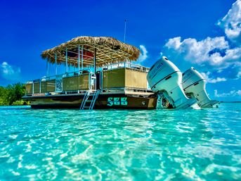 Thatched-roof pontoon boat with twin outboard motors and swim ladder floating in crystal-clear turquoise tropical lagoon under a bright blue sky.