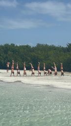 Playful group doing handstands on a sandy, mangrove-lined beach with clear shallow turquoise water and a sunny blue sky.