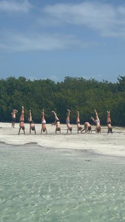Playful group doing handstands on a sandy, mangrove-lined beach with clear shallow turquoise water and a sunny blue sky.