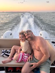 Smiling man and young girl on a boat bench wrapped in towels during a sunset cruise, with twin outboard motors and a foamy wake trailing across open water