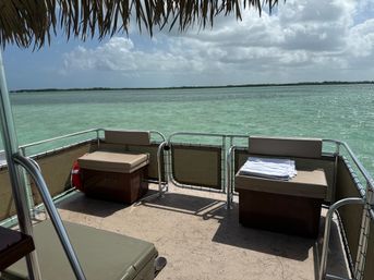 Shaded pontoon boat deck with cushioned storage benches and folded towels overlooking turquoise tropical lagoon and low mangrove-lined horizon under a partly cloudy sky