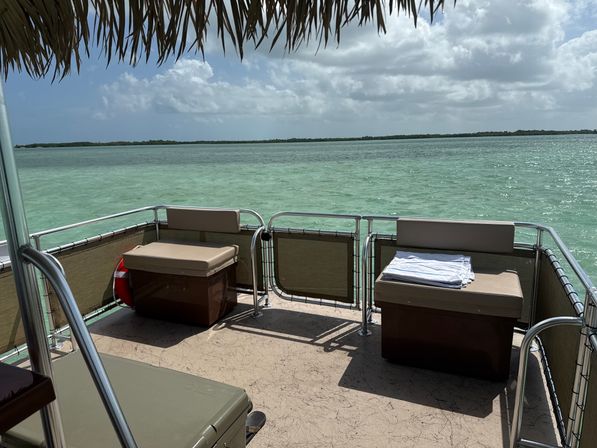 Shaded pontoon boat deck with cushioned storage benches and folded towels overlooking turquoise tropical lagoon and low mangrove-lined horizon under a partly cloudy sky