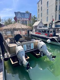 Coastal marina dock with a thatched-roof pontoon boat featuring twin white outboard motors over clear turquoise water, a nearby pilot boat tied to the pier, palm trees and a large marina sign under a sunny blue sky.
