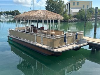 Tiki-style pontoon boat with thatched roof moored at a dock in a calm turquoise marina, boats and a boathouse visible on the palm-lined shoreline