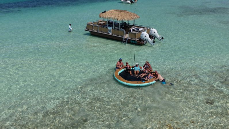 Aerial view of people relaxing on a round floating lounge in shallow turquoise tropical water beside a thatched-roof pontoon boat at a sunny sandbar