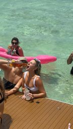 Sunlit dock party in clear turquoise water — a woman in a white bikini leans on the dock as a friend pours a drink from a bottle into her mouth, nearby friends float on a pink inflatable and hold cups.