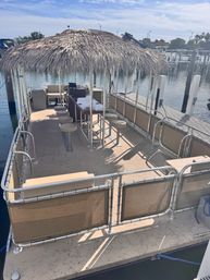 Sunlit pontoon boat with a thatched tiki-hut canopy, central bar with stools and cushioned benches, docked at a calm marina with boats and pilings in the background