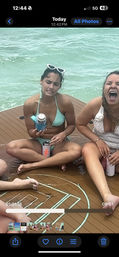 Two women in swimsuits sitting on a boat deck over turquoise ocean water — one in a mint bikini with white sunglasses awkwardly holding a canned drink, the other laughing loudly while holding a tumbler.