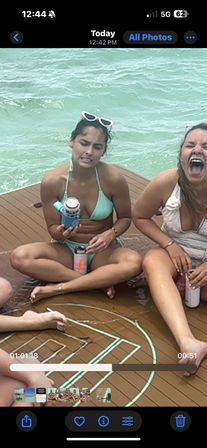 Two women in swimsuits sitting on a boat deck over turquoise ocean water — one in a mint bikini with white sunglasses awkwardly holding a canned drink, the other laughing loudly while holding a tumbler.
