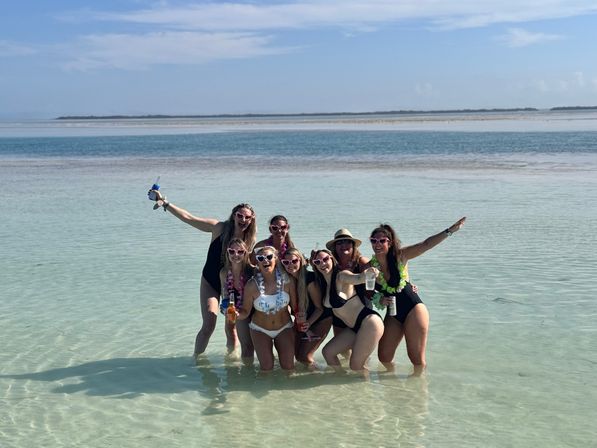 Eight friends in swimsuits and heart-shaped sunglasses posing in clear shallow turquoise water at a sunny tropical beach with a sandy island horizon — celebratory vacation vibe.