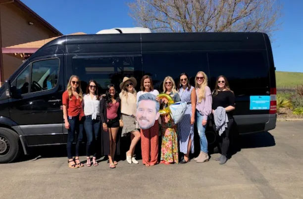 Smiling group of women posing in front of a black party van on a sunny day near rolling vineyard hills, holding balloons and a large face cutout.