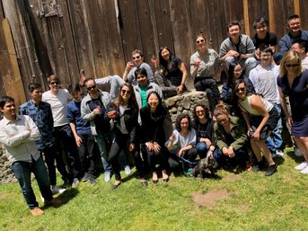 Large mixed group of people posing on a sunny lawn in front of a rustic wooden barn wall; some hold wine glasses, others sit on a stone ledge, and a small dog on a leash sits front and center.