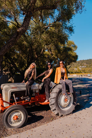 Three fashionable women posing on a vintage tractor under sunlit oak trees on a sunny rural road with blue sky and rolling hills