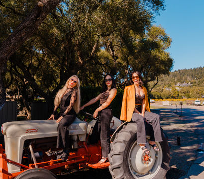 Three fashionable women posing on a vintage tractor under sunlit oak trees on a sunny rural road with blue sky and rolling hills