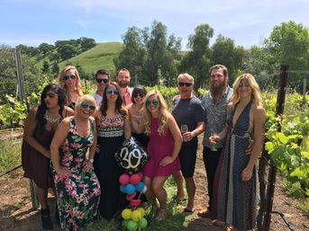 Group of friends posing in a sunny vineyard among grapevines and rolling green hills, holding wine glasses and a colorful balloon cluster topped with a '30' foil balloon.