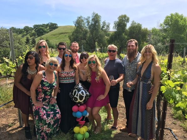 Group of friends posing in a sunny vineyard among grapevines and rolling green hills, holding wine glasses and a colorful balloon cluster topped with a '30' foil balloon.