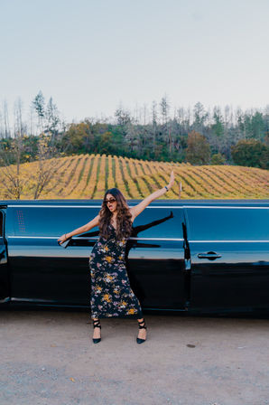 Person in a floral dress and sunglasses striking a playful pose out of a black stretch limousine with golden vineyard rows and tree-covered hillside in the background.