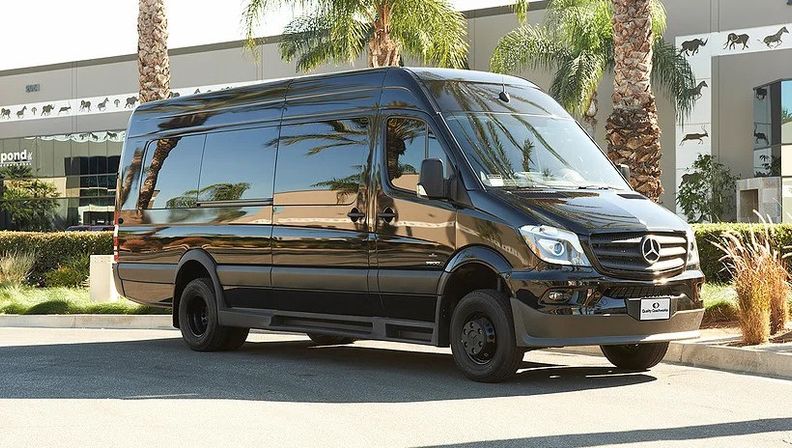 Sleek black Mercedes Sprinter van parked on a sunny palm‑lined commercial street, modern glass building and palm reflections gleaming on its glossy side.