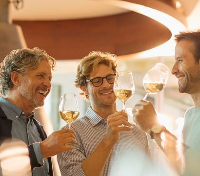Three friends clinking white-wine glasses in a warm-lit modern bar, smiling and toasting during a relaxed wine tasting.