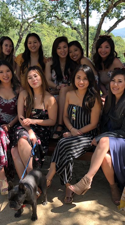 Group of friends in colorful summer dresses posing on picnic benches under oak trees with a sunny hillside vineyard and valley view, a French bulldog on a leash in front.