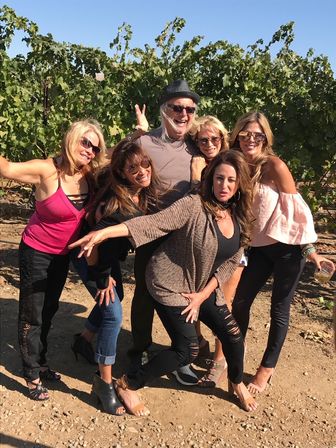 Six friends laughing and posing in a sunny vineyard with grapevines behind them, wearing sunglasses and casual summer outfits for an outdoor wine-tasting vibe.