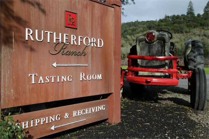 Wooden ranch tasting room sign with directional arrows beside a vintage red tractor on gravel, set against green hills and trees