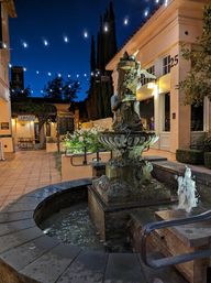 Ornate stone fountain in a tiled outdoor shopping courtyard at dusk, cozy twinkling string lights overhead and warm shopfronts nearby.